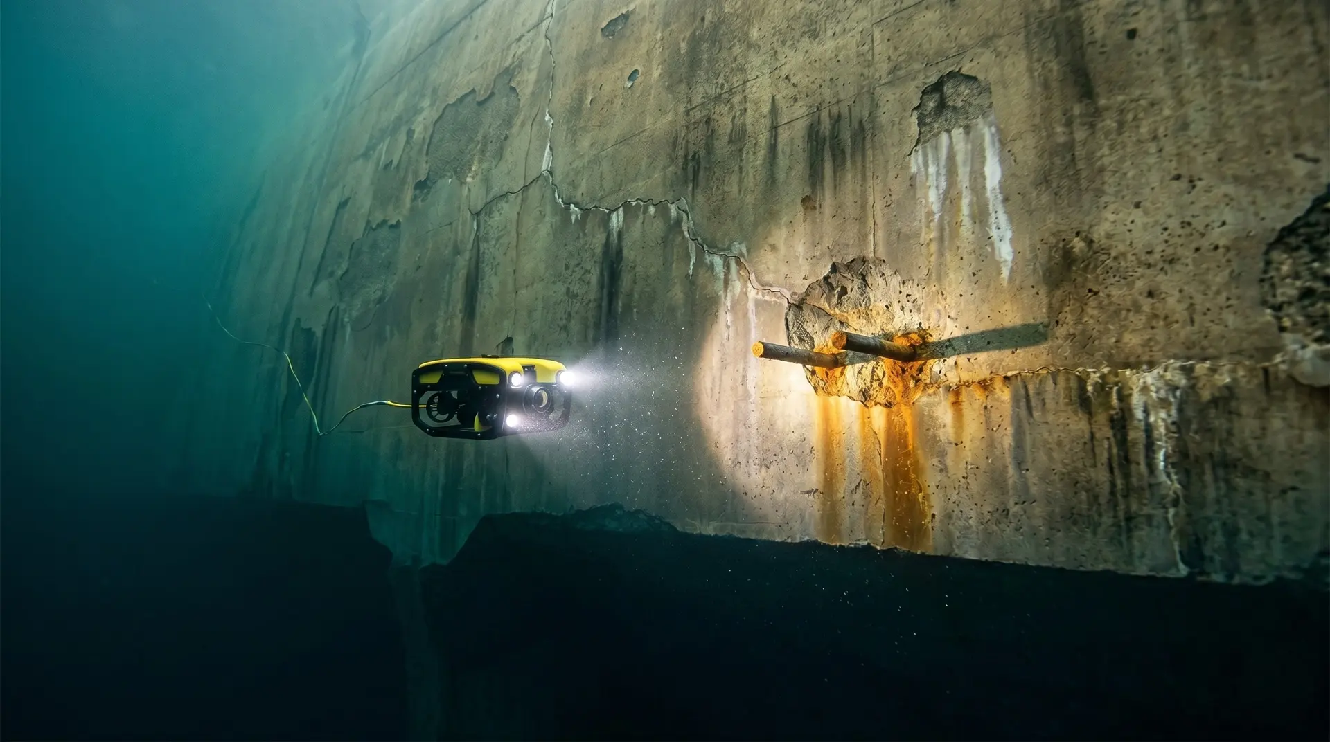 Industrial inspection ROV with twin LED headlights hovering in turbid teal-green reservoir water, illuminating a corrosion-induced spalled zone with exposed corroded rebar stubs on the upstream face of an aging concrete gravity dam
